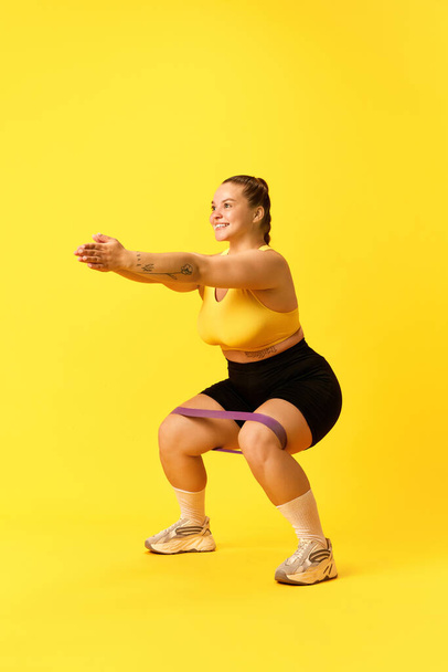 Full length portrait of young chubby sports woman in tracksuit squats with sport elastic bands for legs over yellow background. Concept of sport, hobby, healthy lifestyle, workout, training. ad - Photo, Image