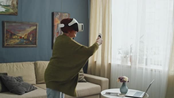 Medium full shot van senior vrouw dragen VR bril en houden bewegingssensoren spelen videospelletjes in het midden van de woonkamer overdag - Video