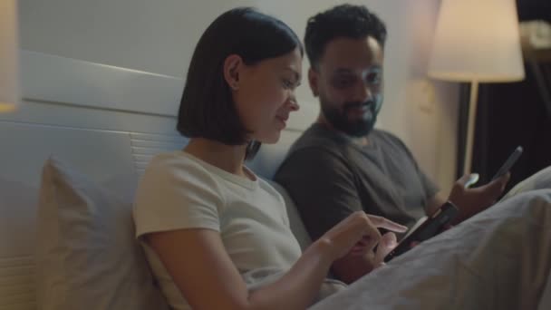 若い家族カップルは夜にベッドに座って,寝る前にスマートフォンでソーシャルメディアについて話し合います - 映像、動画