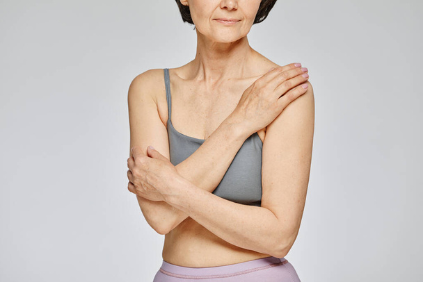 Minimal cropped shot of mature woman wearing neutral underwear against grey background, covering - Photo, Image