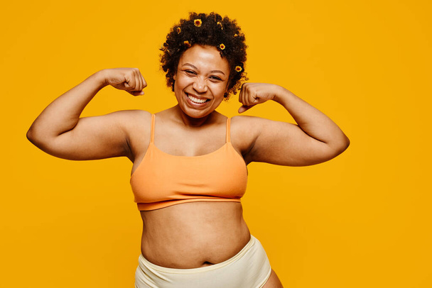 Retrato de cintura hacia arriba de una poderosa mujer negra sonriendo felizmente mientras posa sobre un vibrante fondo amarillo, positividad corporal - Foto, Imagen