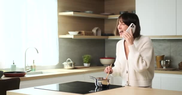 Een multitasking blanke vrouw jonglert met een telefoongesprek tijdens het koken in de keuken. Haar vermogen om beide taken efficiënt te beheren weerspiegelt haar bekwaamheid en aanpassingsvermogen in een druk huishouden. - Video
