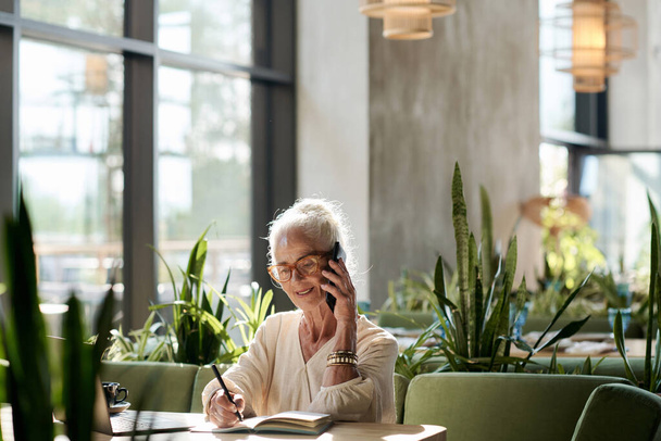 Femme âgée ayant une conversation sur son téléphone portable et prenant des notes dans son bloc-notes pendant son travail dans un café - Photo, image