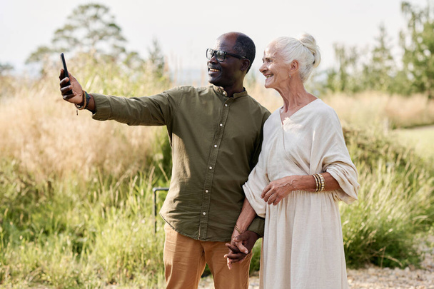 Senior couple making selfie portrait on smartphone while walking outdoors - Photo, Image