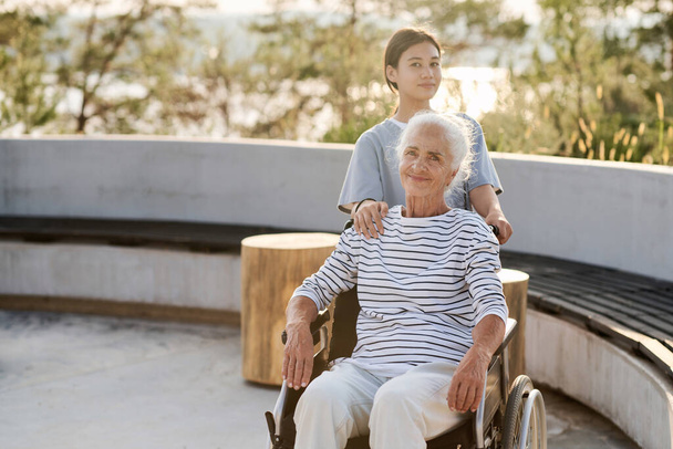 Porträt einer Seniorin, die im Rollstuhl sitzt und beim Gehen mit einer Krankenschwester in die Kamera schaut - Foto, Bild