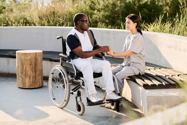 Jonge verzorger zit op de bank en praat met oudere man met een handicap tijdens hun wandeling buiten - Foto, afbeelding