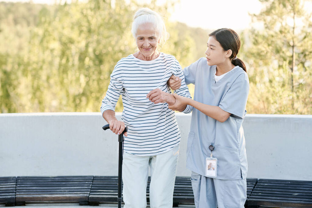 Caregiver helping senior woman to walk with stick during their walk outdoors - Photo, Image