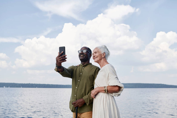 Multietnico felice coppia anziana fare selfie sul telefono cellulare contro il lago all'aperto - Foto, immagini