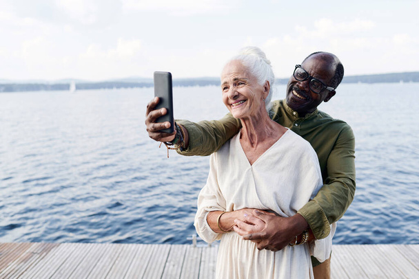 szczęśliwy starszy człowiek robi selfie z jego żoną podczas gdy oni stoją na molo nad jeziorem - Zdjęcie, obraz