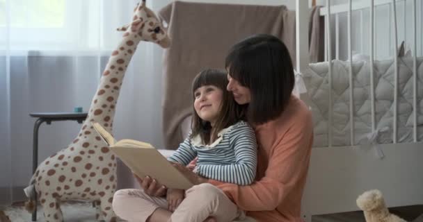 In de vrolijke kinderkamer lezen een vrolijke moeder en haar dochter samen een boek. Hun gedeelde enthousiasme voor lezen vult de kamer met warmte en liefde voor verhalen vertellen. - Video