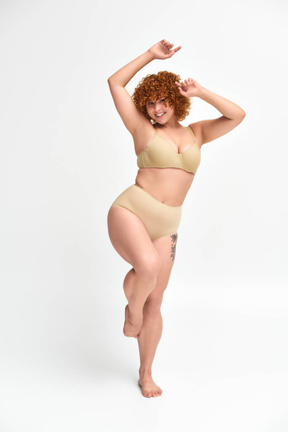 full length of jolly redhead woman with curvy body posing on one leg with raised hands on white - Photo, Image