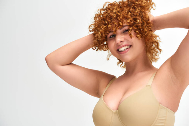 cheerful young woman with curvy breasts touching red wavy hair on white backdrop, plus size beauty - Photo, Image