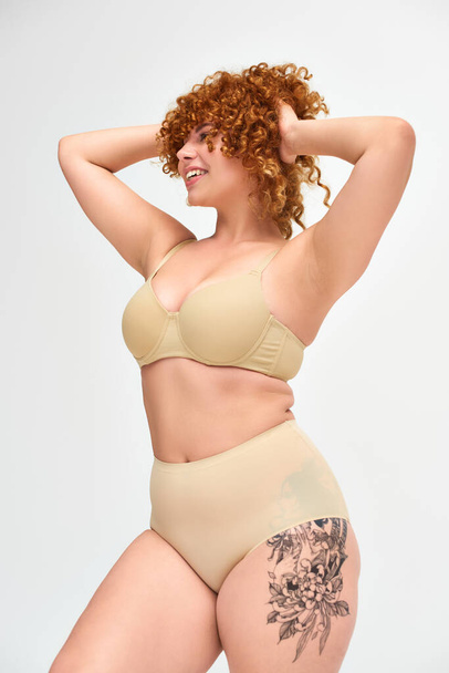 tattooed curvy woman with red curly hair posing in beige lingerie with hands behind head on white - Photo, Image