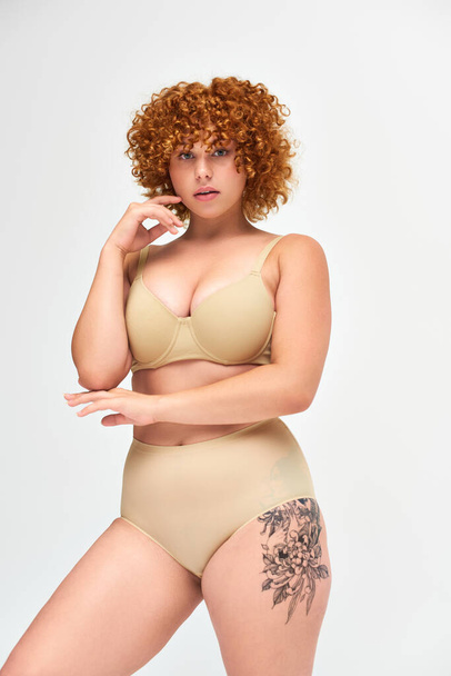 sensuale tatuato plus size donna in biancheria intima taupe con capelli rossi ondulati guardando la fotocamera su bianco - Foto, immagini