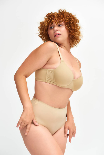 body positive redhead and curly woman in taupe underwear looking away on white, self-expression - Photo, Image