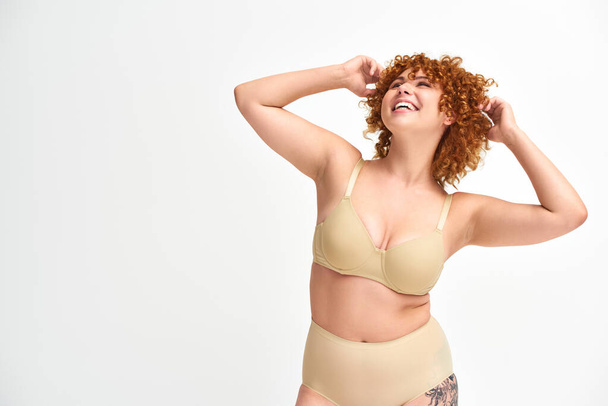 overjoyed and redhead plus size woman in beige underwear smiling and looking away on white - Photo, Image