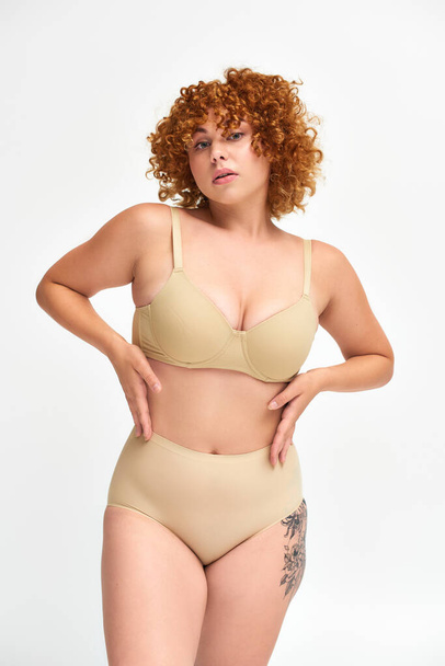 sensual redhead plus size woman in beige lingerie posing with hands on waist on white, self-esteem - Photo, Image