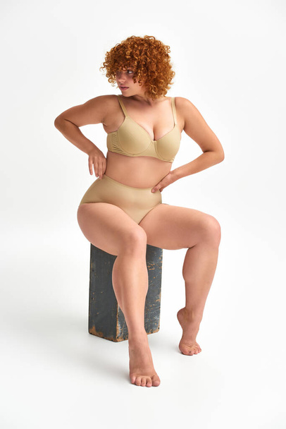 redhead and curvaceous woman in beige lingerie sitting on wooden box and looking away on white - Photo, Image