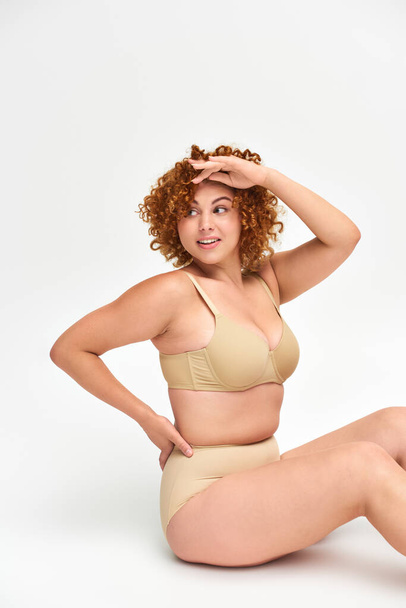 smiling redhead plus size woman in beige lingerie sitting with hand on hip and looking away on white - Photo, Image