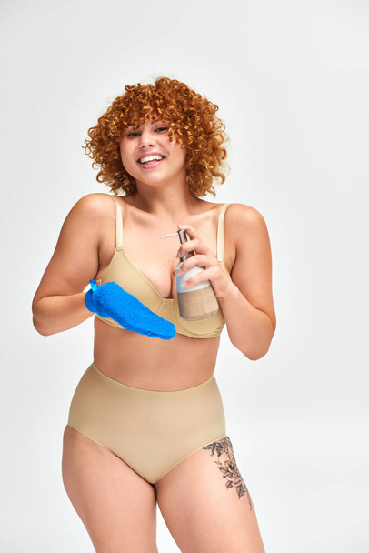 šťastná zrzka plus velikost žena ve spodním prádle se sprchovým gel dávkovač a koupací rukavice na bílém - Fotografie, Obrázek