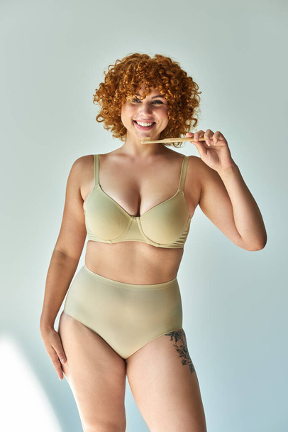 joyful redhead plus size woman in beige underwear holding toothbrush and looking at camera on beige - Photo, Image
