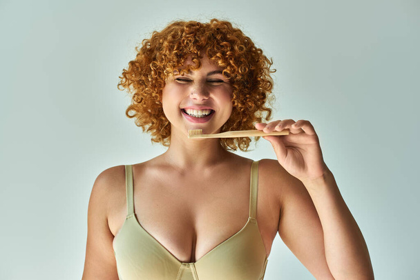 cheerful redhead and curvy woman with toothbrush smiling with closed eyes on beige backdrop - Photo, Image