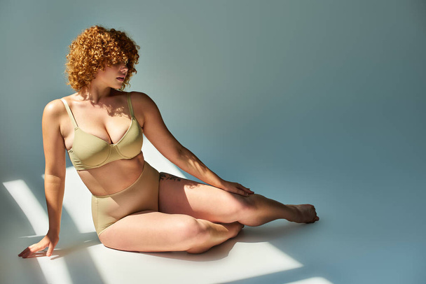 alluring and redhead plus size woman in beige lingerie sitting in sunshine on grey backdrop - Фото, изображение