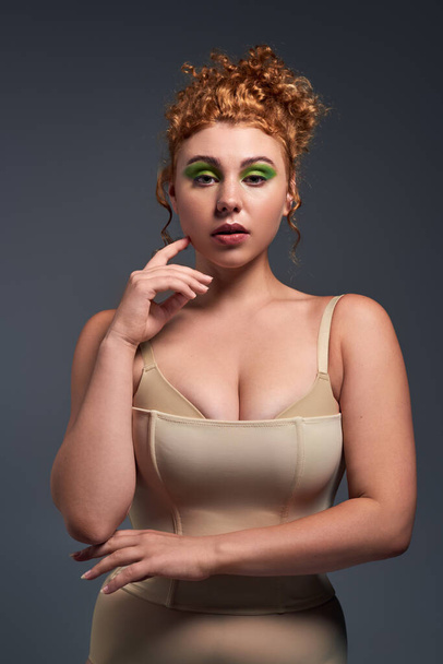 portrait of young redhead and curvy woman in bra and corset looking at camera on dark grey backdrop - Photo, Image