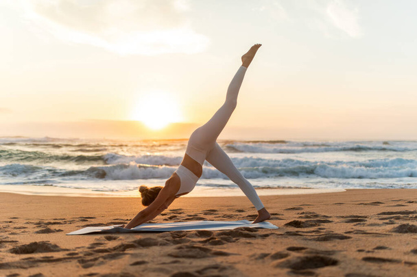 Młoda kobieta w odzieży sportowej ćwiczy jogę z podniesioną nogą na świeżym powietrzu, ćwiczy na plaży morskiej o zachodzie słońca, widok z boku, pełna długość. Pani rozciągająca się na zewnątrz - Zdjęcie, obraz