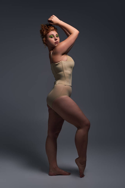 full length of redhead plus size model in beige lingerie on dark grey backdrop, expressive pose - Photo, Image