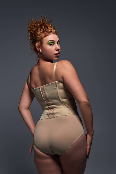 back view of curvaceous redhead woman in beige lingerie looking at camera on dark grey backdrop - Photo, Image