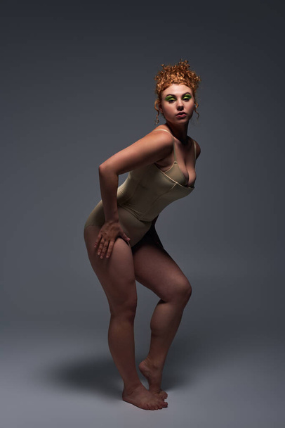 volledige lengte van jonge roodharige plus size vrouw in beige ondergoed in expressieve pose op donkergrijs - Foto, afbeelding
