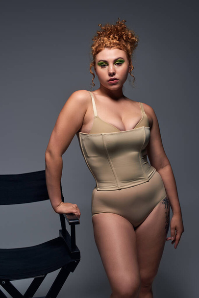 redhead and thoughtful plus size model in beige underwear standing near chair on dark grey backdrop - Photo, Image