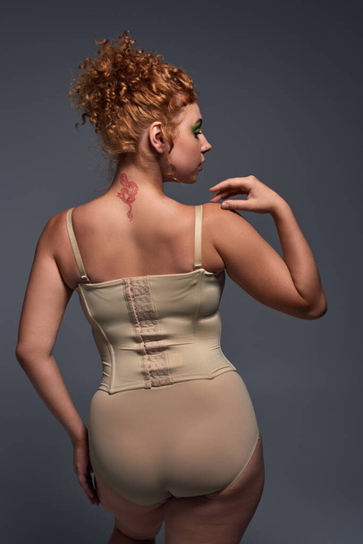 back view of redhead plus size woman in beige underwear on dark grey backdrop, body confidence - Photo, Image