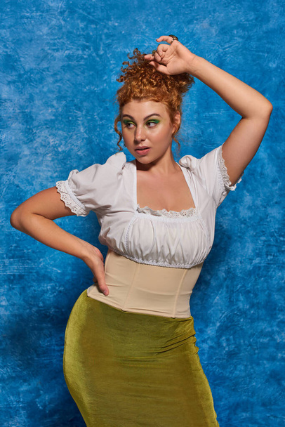 young redhead plus size woman in stylish clothes posing with hand on hip on blue textured backdrop - Photo, Image