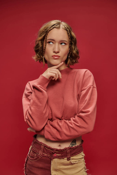 задумчивая стильная и молодая женщина в лоскутных штанах и обрезанный длинный рукав позирует на красном фоне - Фото, изображение