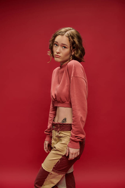 mirada inocente, mujer joven en pantalones patchwork y manga larga recortada posando sobre fondo rojo - Foto, imagen
