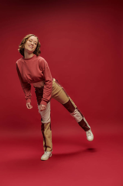 personal style, young positive girl in patchwork pants and cropped long sleeve posing, red backdrop - Photo, image