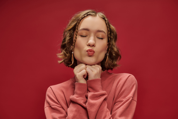 portrait of young pretty woman with short wavy hair and braids pouting lips on red, air kiss - Photo, Image