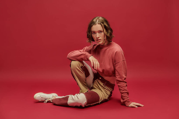 self-expression, young woman in patchwork pants and cropped long sleeve sitting on red backdrop - Photo, Image