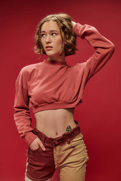 personal style, young pretty woman in patchwork pants and cropped long sleeve posing on red backdrop - Photo, Image