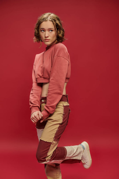 stile giovanile, timida bella donna in pantaloni patchwork e manica lunga ritagliata in posa su sfondo rosso - Foto, immagini
