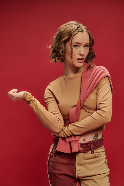 trendige junge Frau in Patchworkhosen posiert im Pullover über abgeschnittenem Langarm vor rotem Hintergrund - Foto, Bild