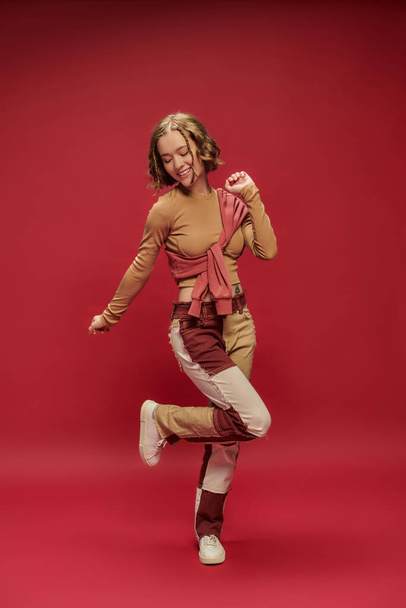 excited young woman in patchwork pants posing in jumper over cropped long sleeve on red backdrop - Photo, Image