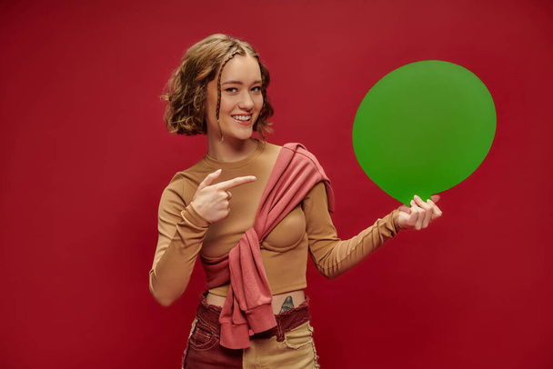 щаслива жінка в клаптикових брюках і перестрибувати над обрізаним довгим рукавом, вказуючи на мовну бульбашку на червоному - Фото, зображення