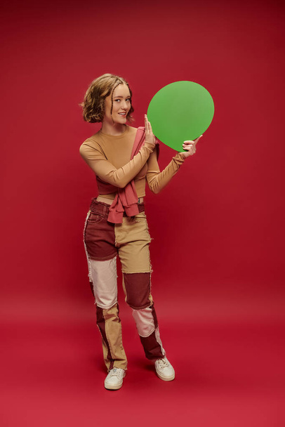 νεαρή γυναίκα σε patchwork παντελόνι και jumper πάνω περικοπεί μακρύ μανίκι δείχνοντας φούσκα ομιλία στο κόκκινο - Φωτογραφία, εικόνα