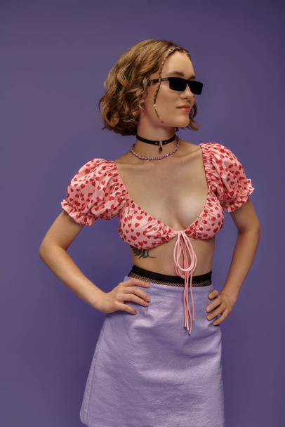 fiduciosa giovane donna in occhiali da sole alla moda e top ritagliato in posa con le mani sui fianchi su viola - Foto, immagini