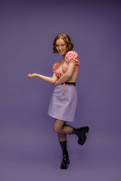 усміхнена молода жінка в обрізаному верху і спідниці позує в чоботях і ажурних колготках на фіолетовому фоні - Фото, зображення