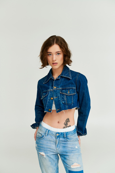 stylová mladá žena s tetováním pózuje v střižené džínové bundě a modré džíny na šedém pozadí - Fotografie, Obrázek