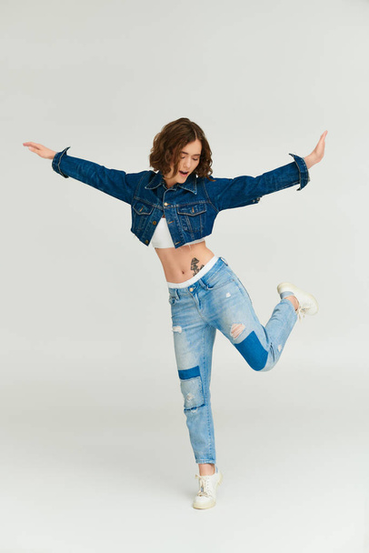 stylish young woman with tattoo in cropped denim jacket and jeans standing on leg on grey background - Photo, Image